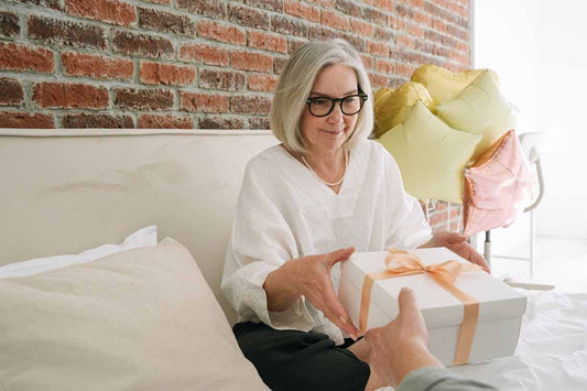 Older woman sitting on a bed receiving a wrapped gift, with balloons in the background.